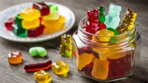 A jar of colorful gummy bears on a wooden table, with some gummies scattered around and a plate of more gummies blurred in the background. The gummies come in various shapes and bright colors like red, yellow, green, and orange, creating an inviting and playful display.